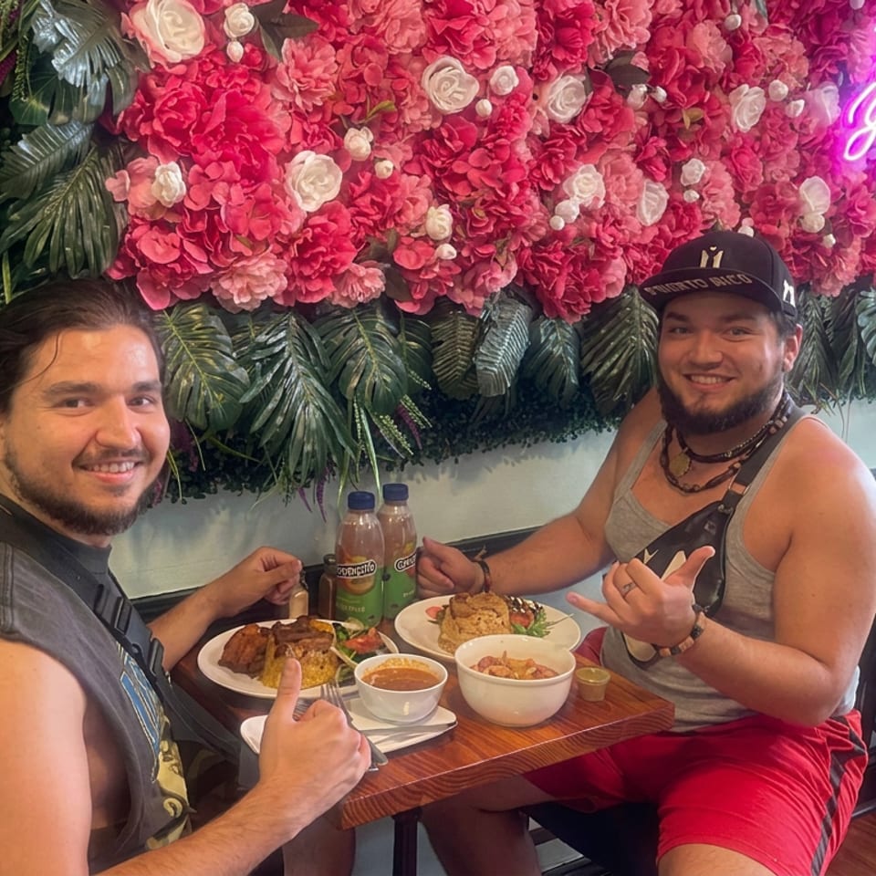 Two smiling men pose over plates of food at a restaurant table beneath a vibrant pink floral wall and neon sign.
