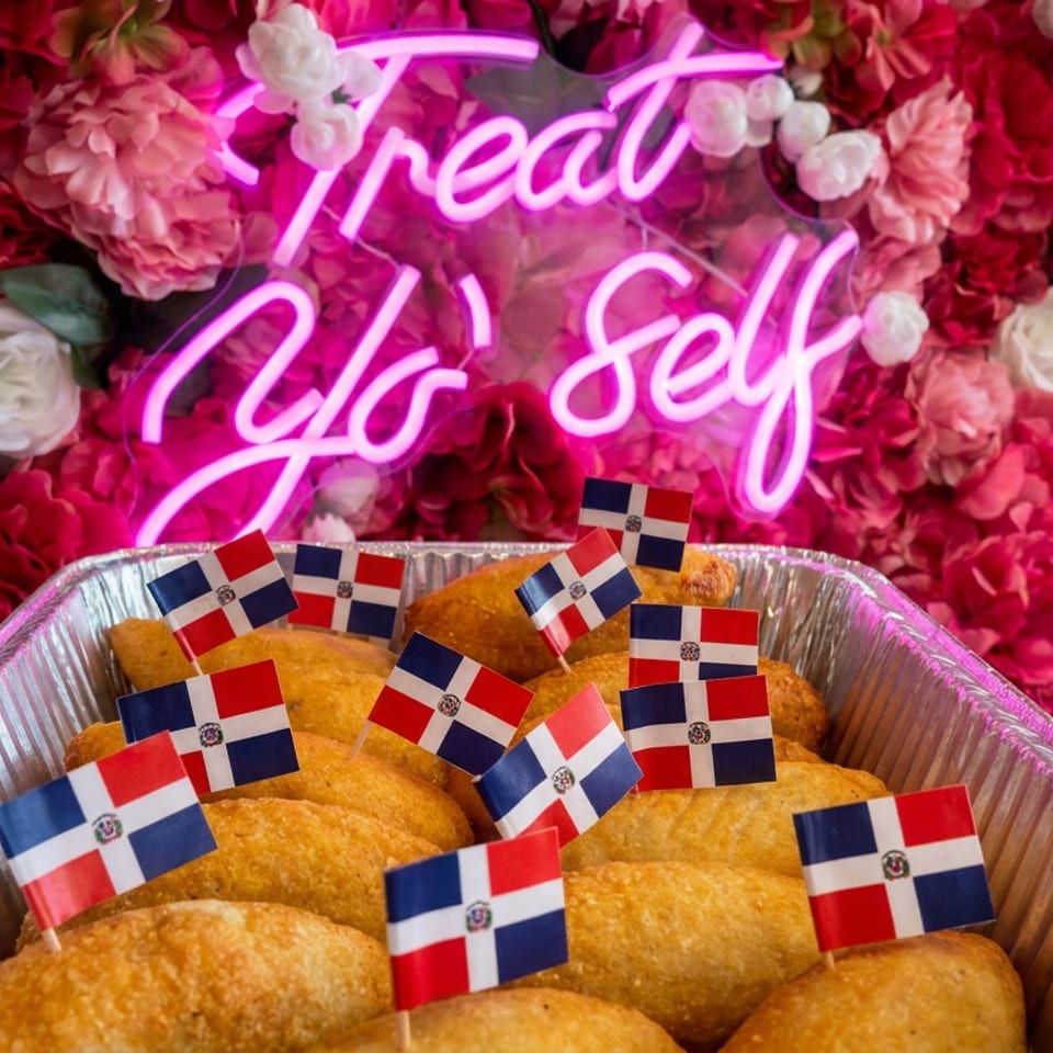 Golden Dominican pastries in a foil tray decorated with Dominican flags against a pink floral wall and neon “Treat Yo’ Self” sign.