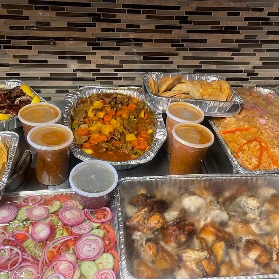 Catering spread displays grilled meat, stewed vegetables, pastries, rice, roasted chicken, salad, sauces, and colorful garnishes in aluminum trays.
