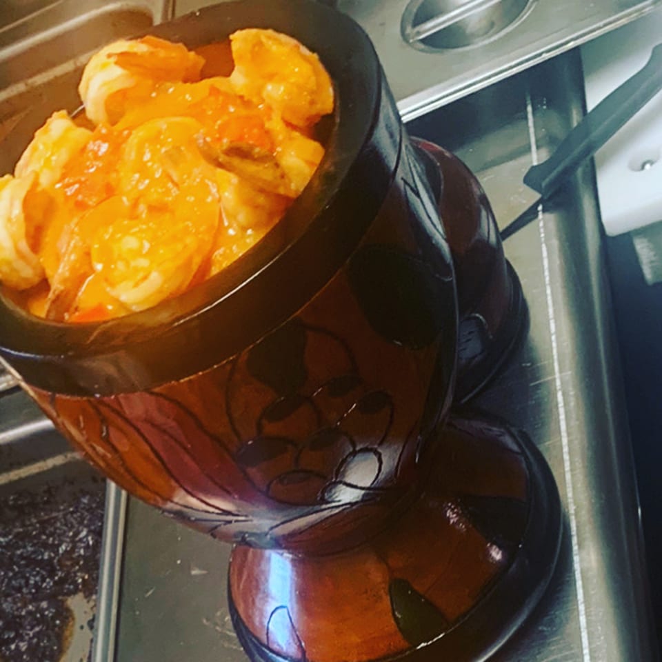 Glazed shrimp served in a dark ceramic bowl beside a stainless-steel buffet counter.
