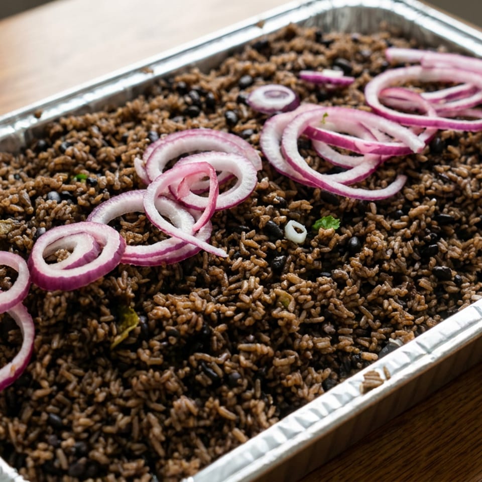 Tray of seasoned rice and black beans garnished with sliced red onions and herbs on a wooden table.